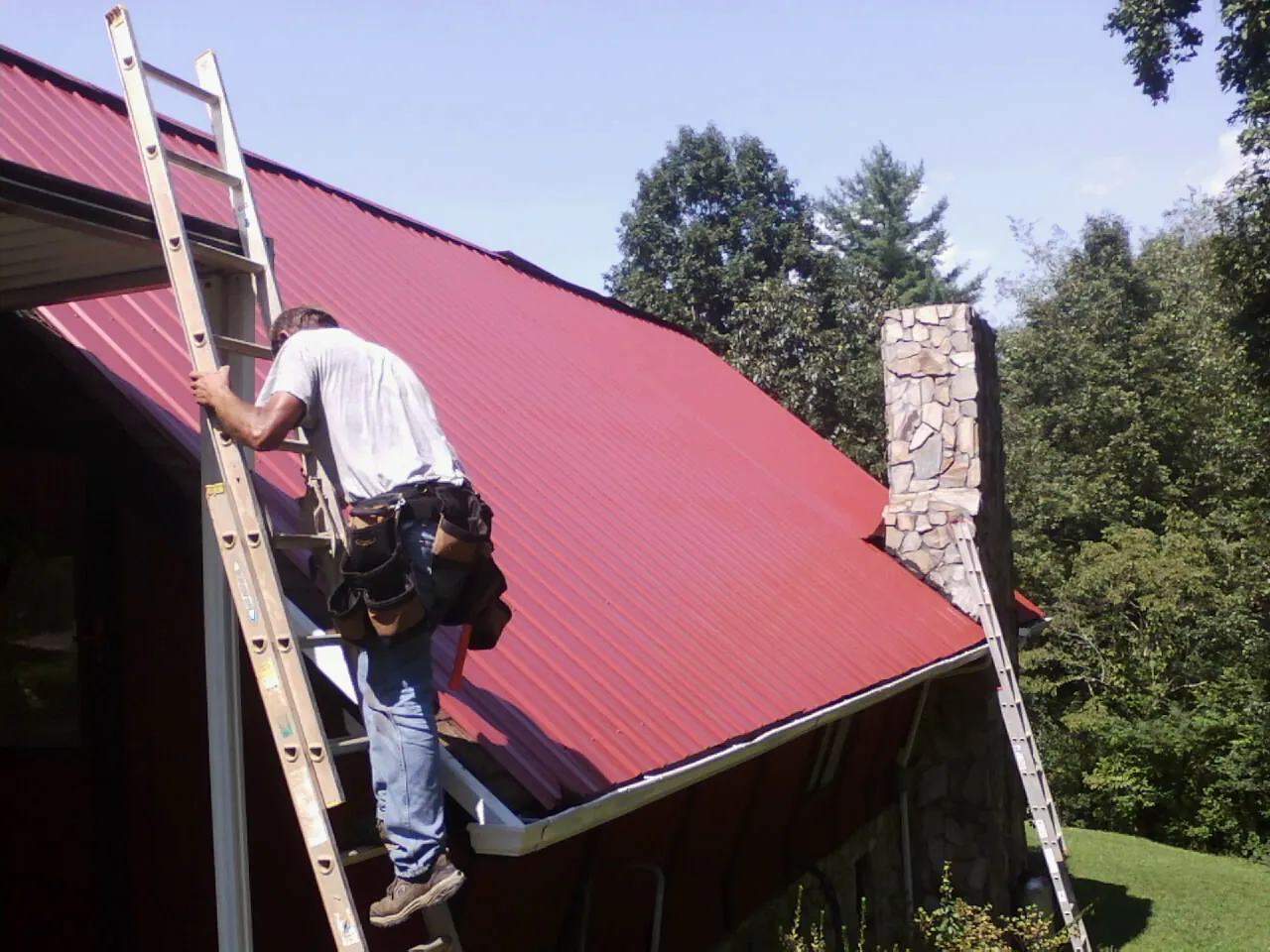 Roofer on a ladder beside a red metal roof and stone chimney — Soffit & Fascia Repair by Roofing Brothers in Locust Dale
